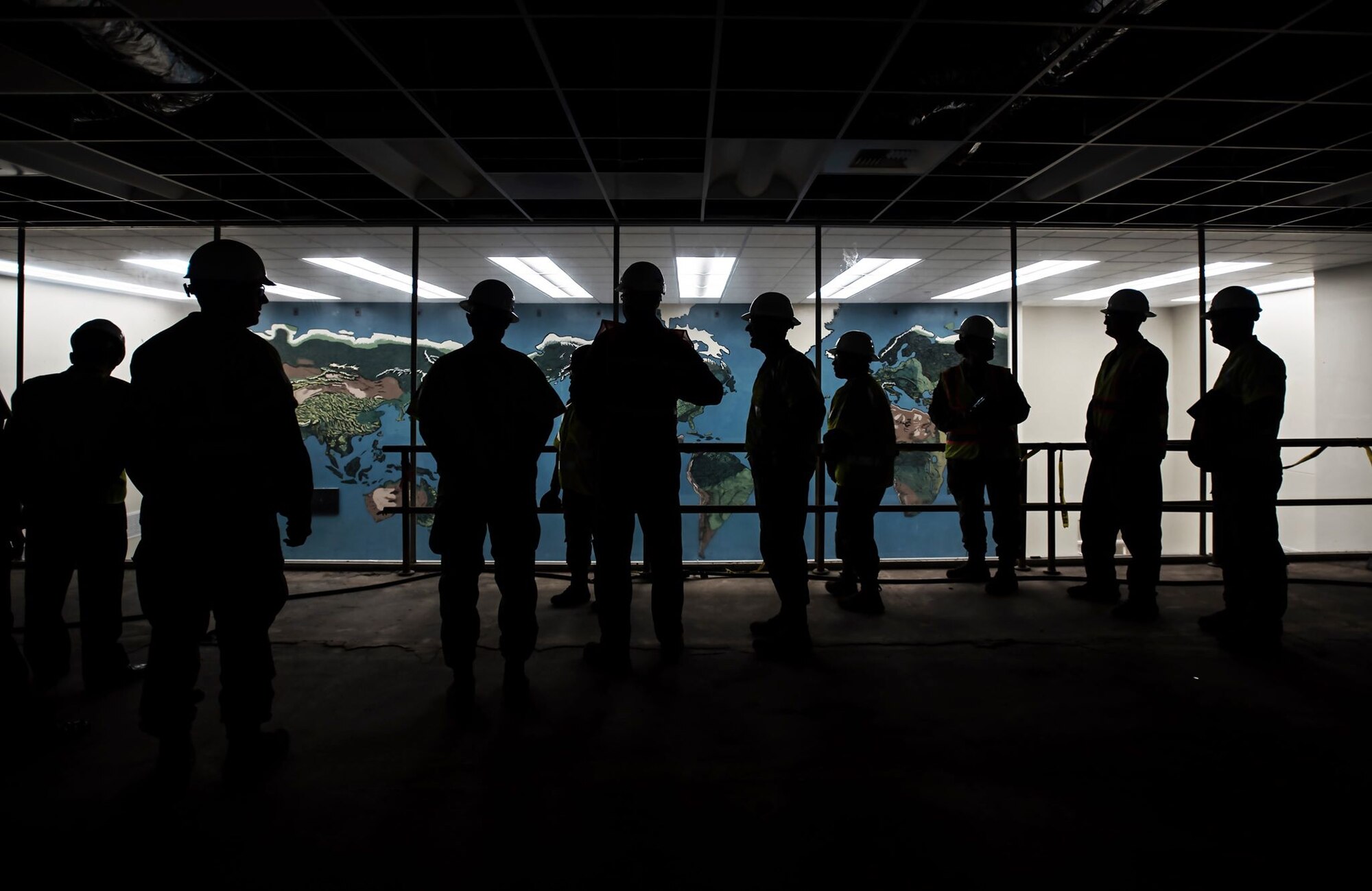A group of military personnel in hard hats are silhouetted against the brightly lit room in front of them with a map of the world on the wall