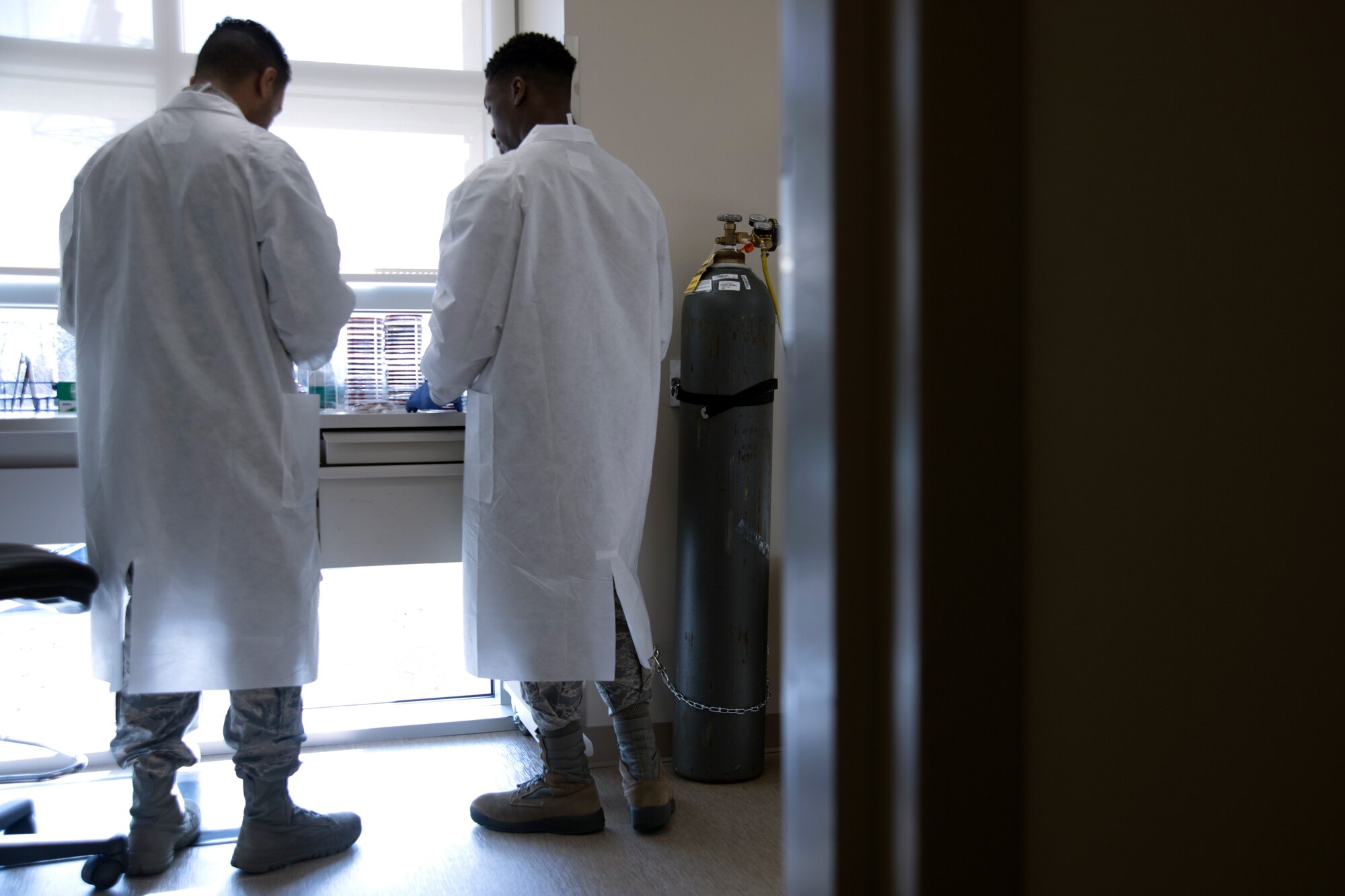 Photo of two Airmen in lab coats facing away to look at a table with medical equipment