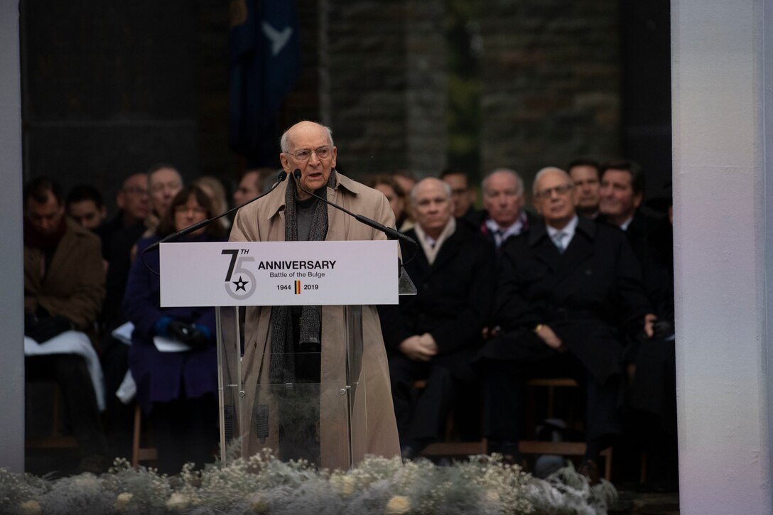 Man speaks at a lectern that bears a sign commemorating the 75th anniversary of the Battle of the Bulge.