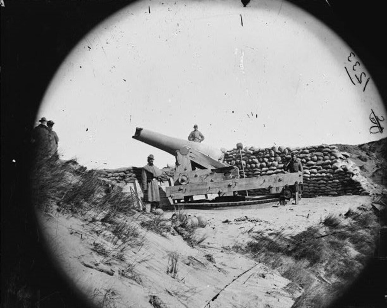 Three men stand beside a large cannon on a beach. Behind it is a pile of sandbags.
