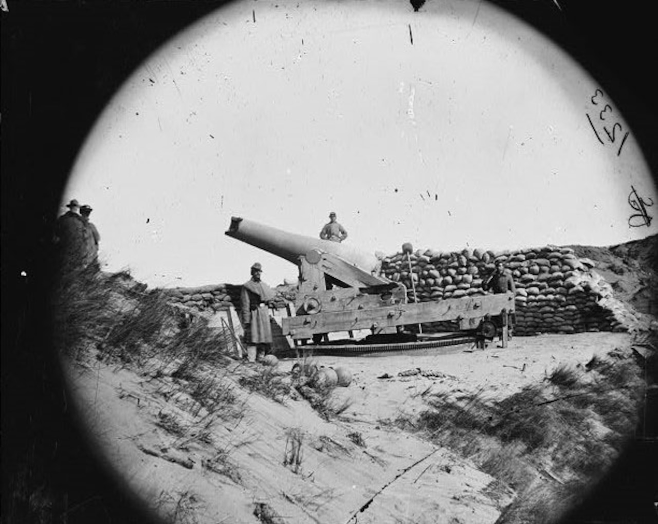 Three men stand beside a large cannon on a beach. Behind it is a pile of sandbags.