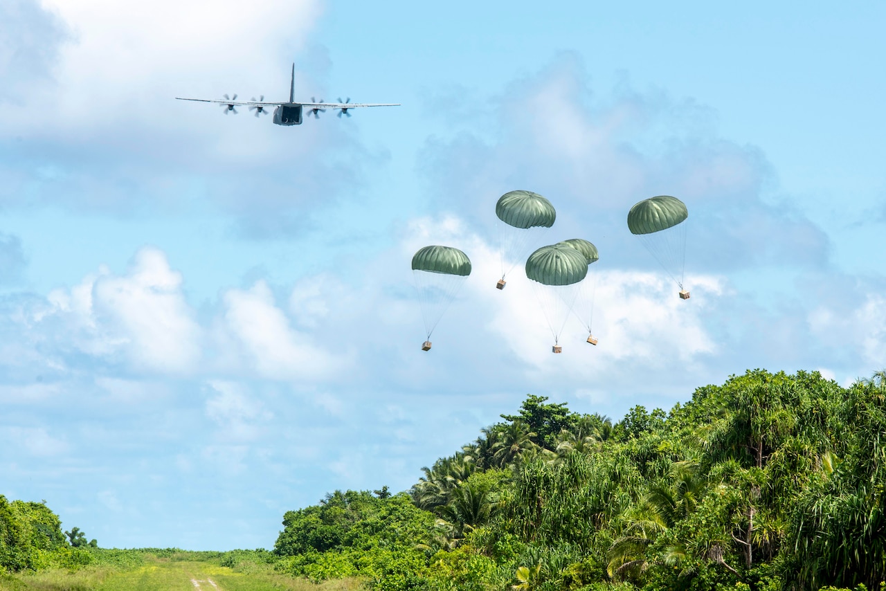 Bundles attached to parachutes descend over a green landscape as an aircraft flies nearby.