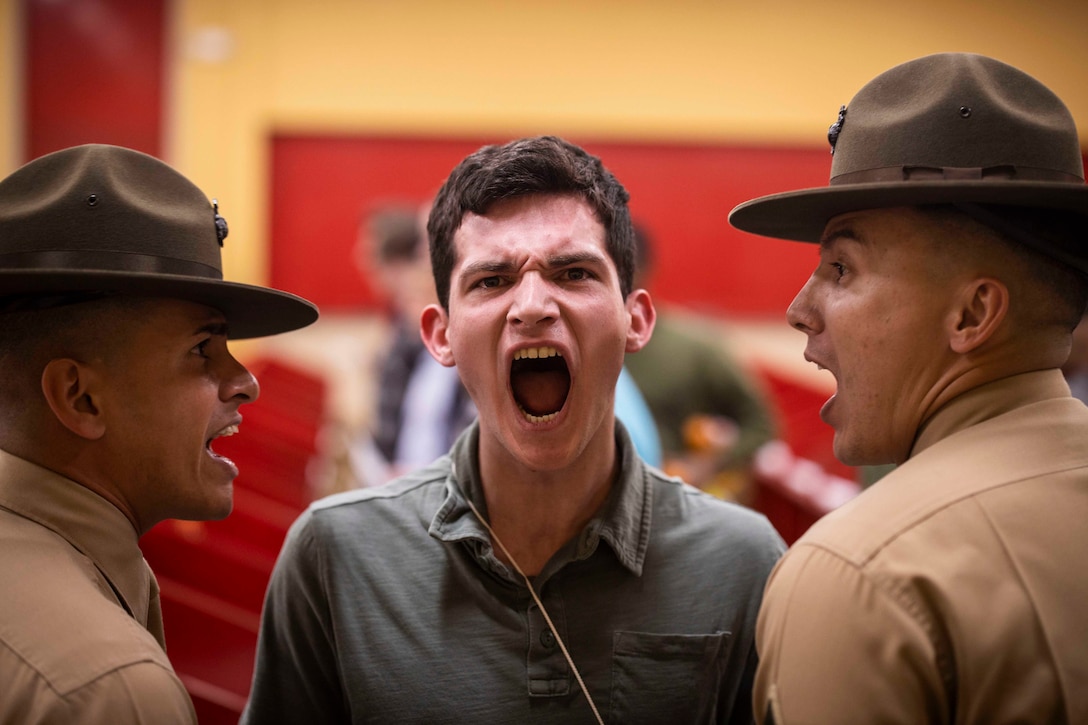 A Marine Corps recruit yells as two drill instructors yell on either side.