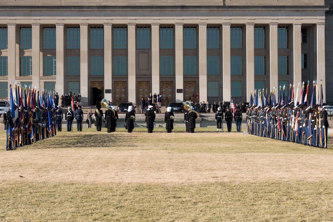 Defense leaders stand on the steps of the Pentagon while two rows of service members stand in the foreground holding flags.