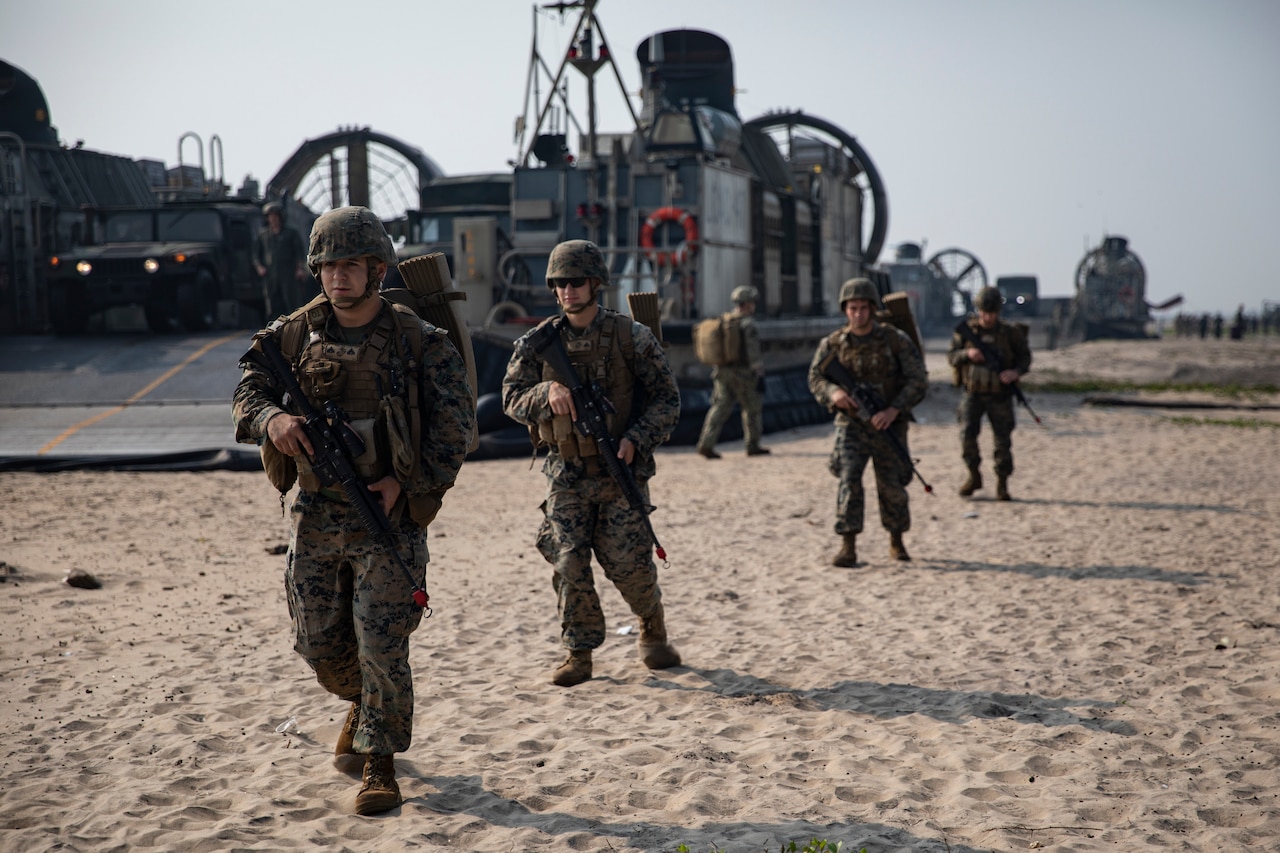 Service members walk across sand and carry rifles.  In the background is a large hovercraft military vehicle.
