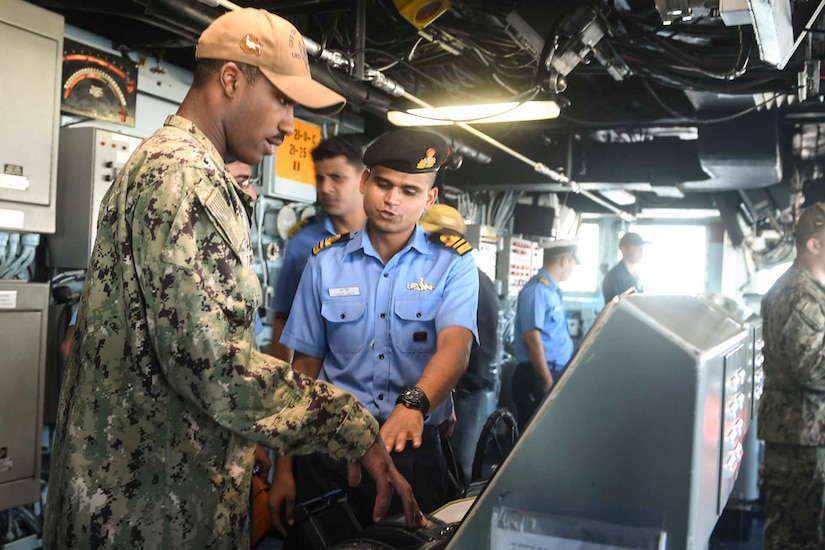 A uniformed service member points at military equipment while another uniformed service member watches.