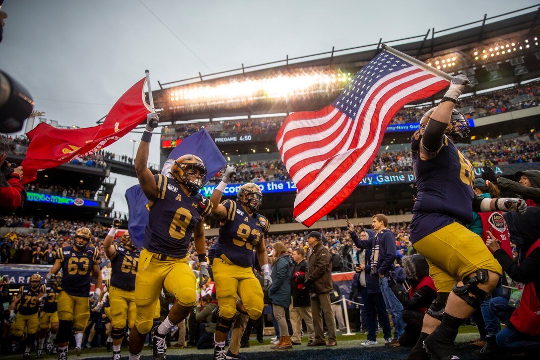 U.S. Naval Academy football players, some waving flags, run onto a field.