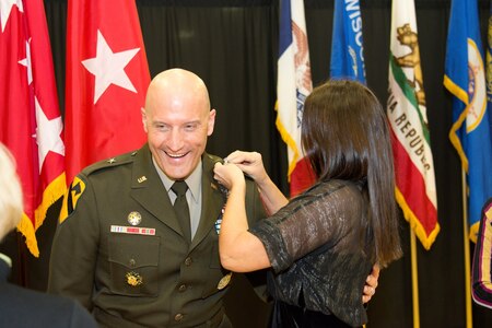 Woman in black dress pinning rank on shoulder of male Soldier in green uniform