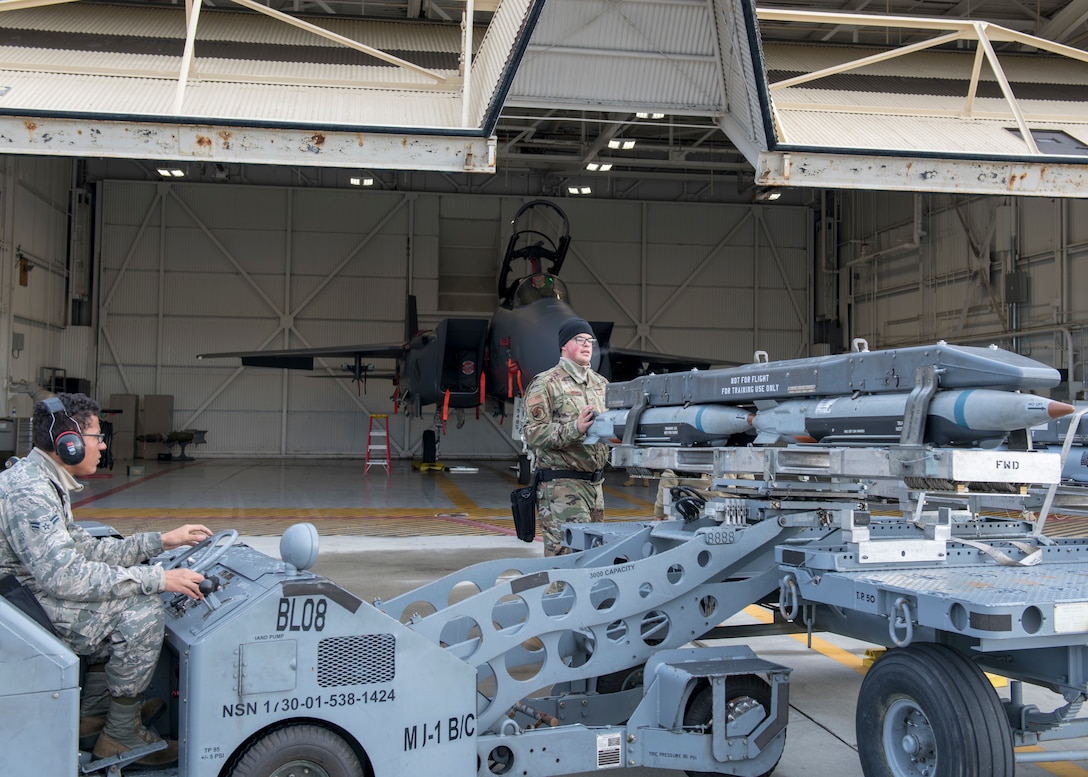 Airman 1st Class and AMU load crew technician work together to load a Bomb Rack Unit 61 on an MJ-1 lift truck