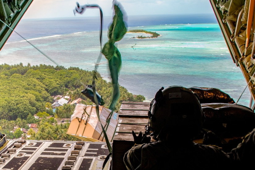 A package attached to a parachute drops out of an open aircraft over an island as an airman watches.