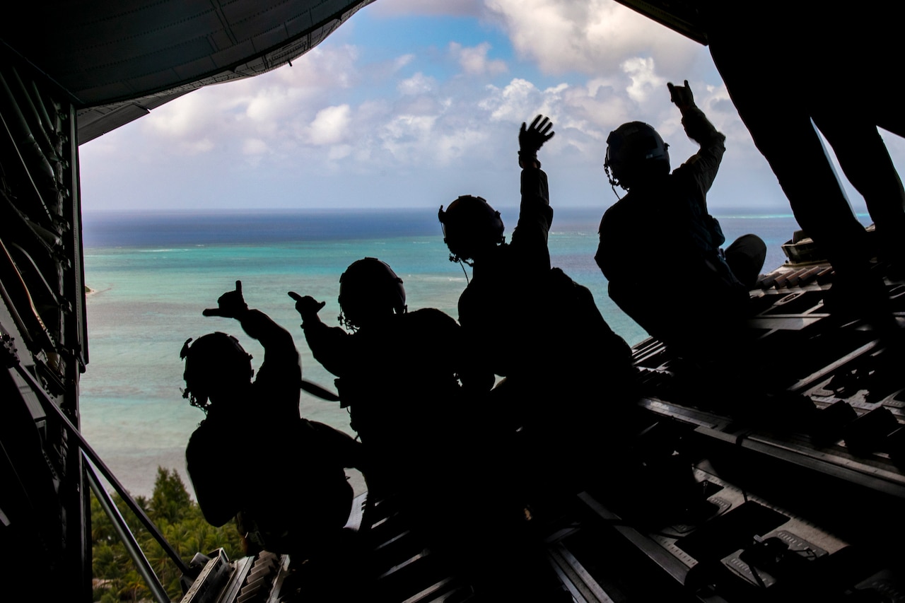 Four airmen wave out of the back of an open aircraft flying over an island.