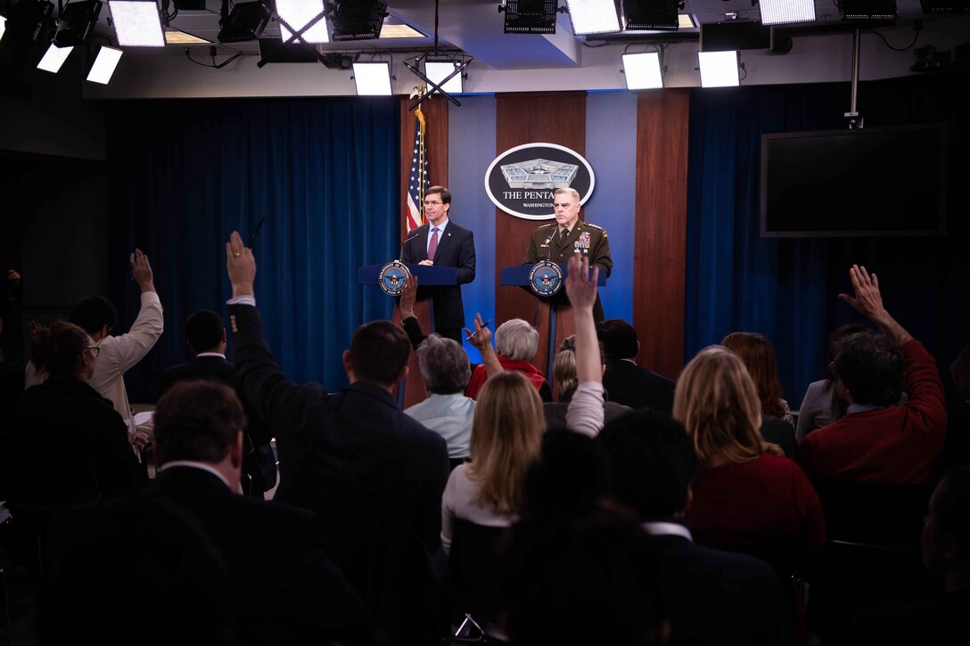 Two men stand behind lecterns; some people in the audience raise their hands.