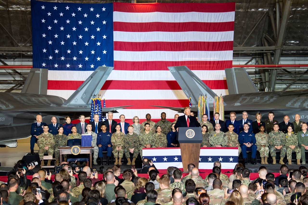 President Donald J. Trump stands on stage with a group of people; a large crowd sits in the audience.