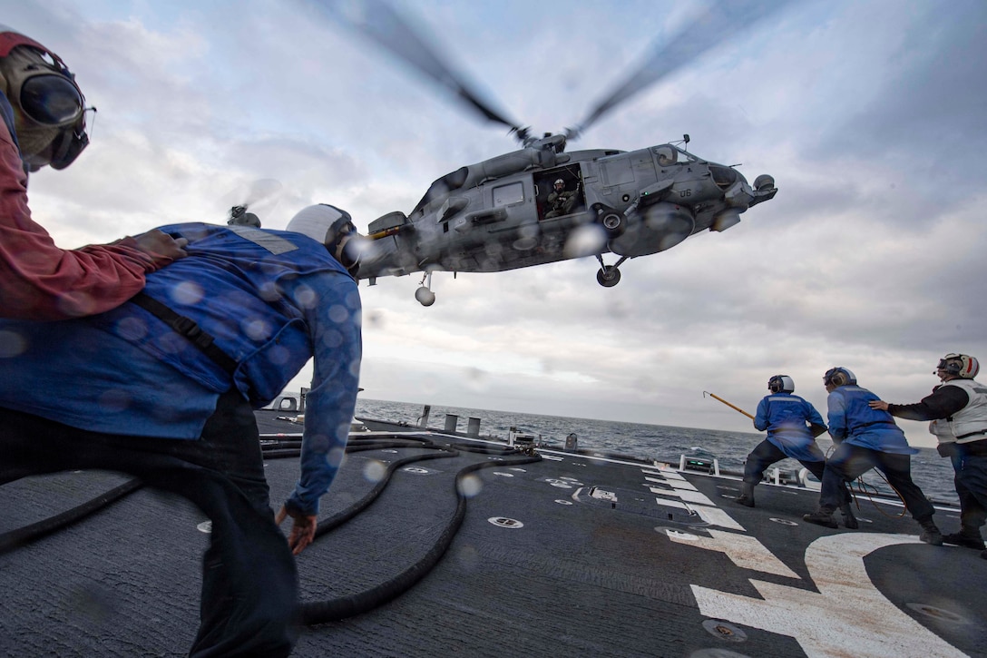 A group of sailors crouch on the flight deck of a military ship as a helicopter hovers above.