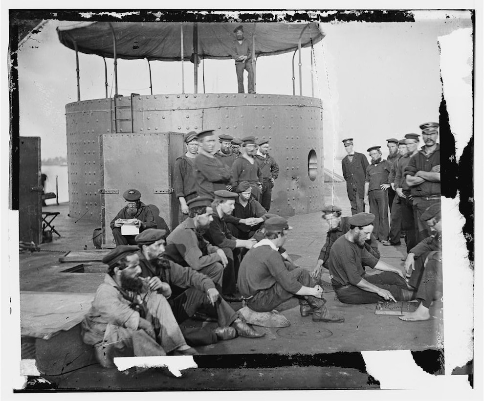 Several Civil War-era sailors sit on the deck of an ironclad warship. A large, round turret is nearby.