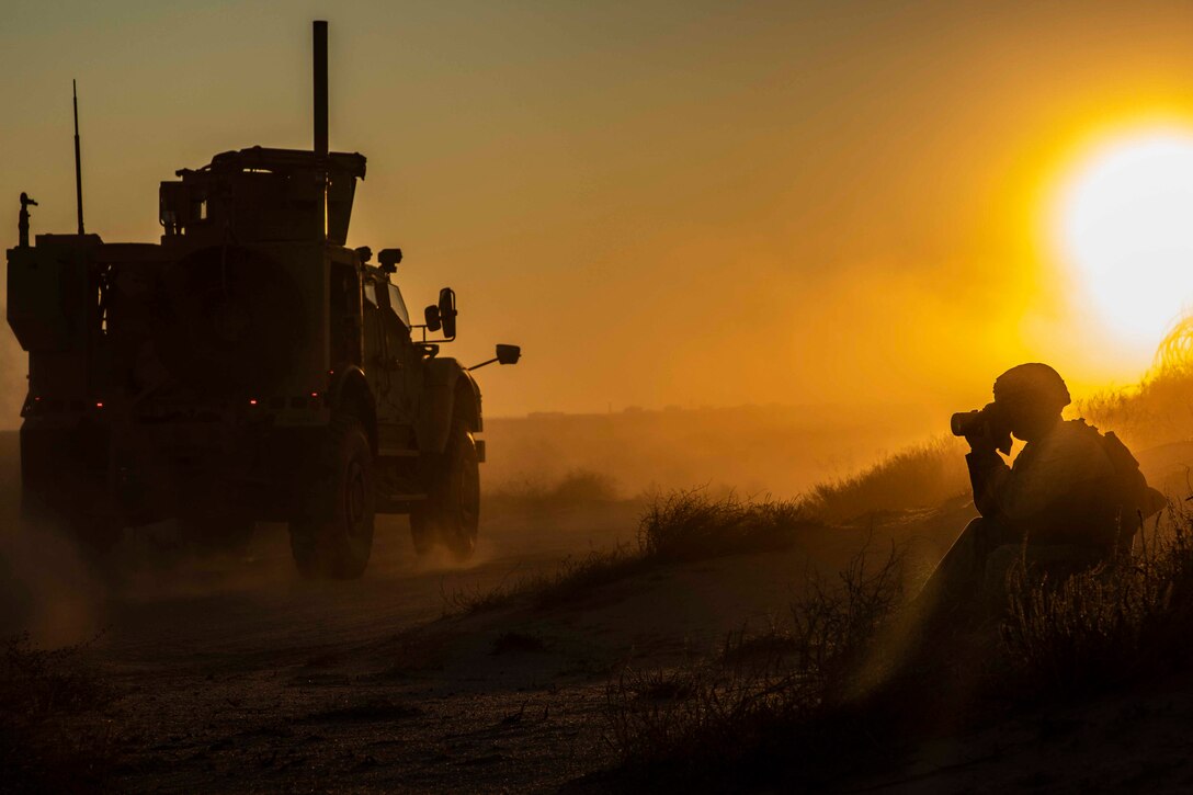 A Marine takes a photo of a moving vehicle as the sun shines in the background.