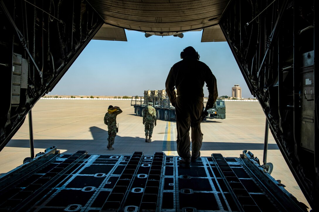 An airman stands in the back of an open aircraft as other troops walk on a flightline.