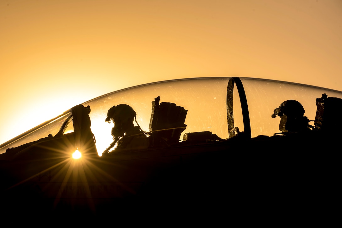 Two airmen, shown in silhouette against an orange sky, sit in a cockpit.