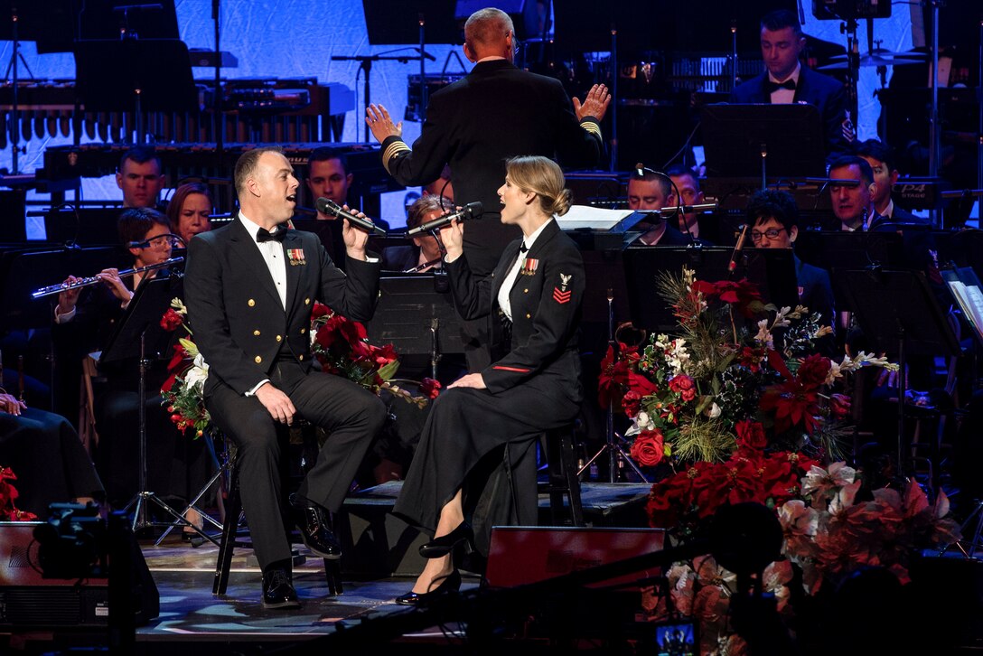 Two sailors on stools sing on a stage, as a band plays behind them.