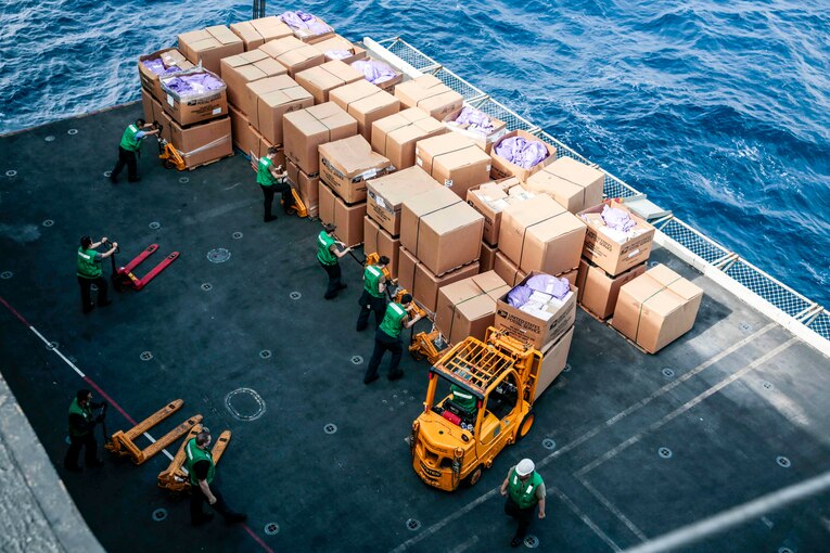 Sailors, seen from overhead, work by large cardboard boxes stacked on the deck of a ship in the water.
