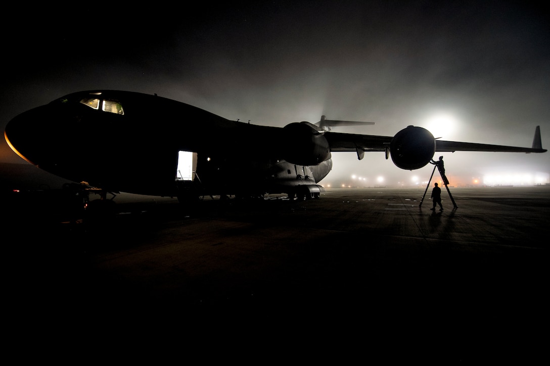 An airman on a ladder, shown in silhouette, inspects an aircraft engine on a dark flightline.