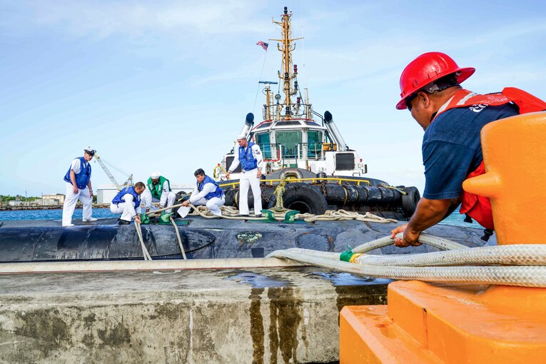 Sailors atop a submarine and one on shore handle lines while mooring the sub.