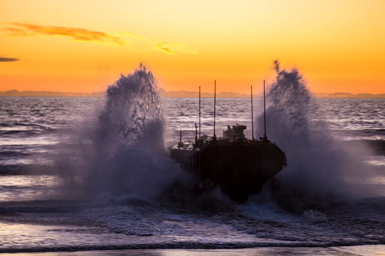 A Marine Corps amphibious vehicle splashes in ocean water, appearing purplish against an orange sky.