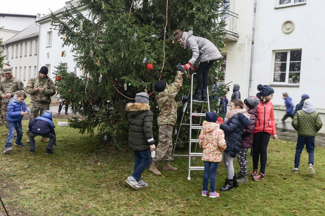 Soldiers and children decorate a Christmas tree outside a white building.