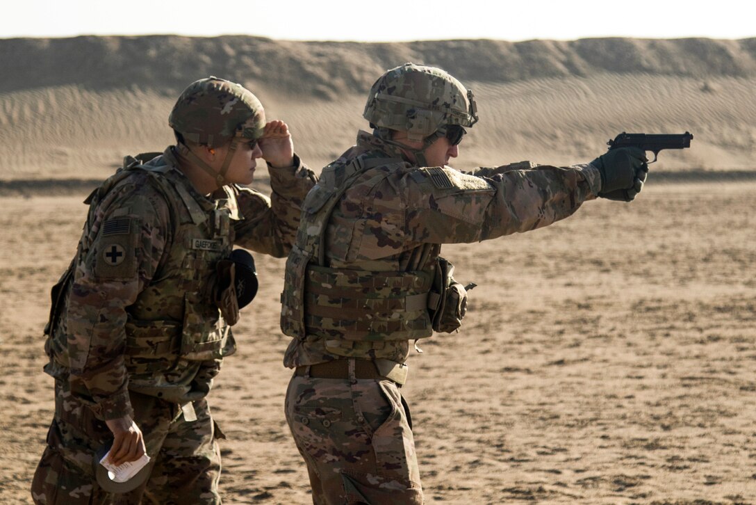 A soldier aims a pistol at an outdoor range as another soldier watches behind him.