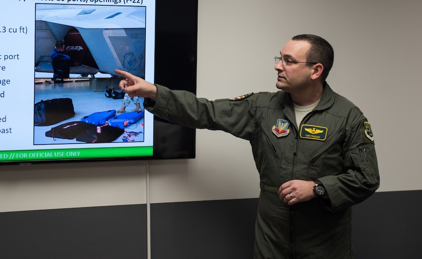 U.S. Air Force Major Brian Bascuzzi, 1st Fighter Wing chief of innovation, points to a slide during the ribbon cutting ceremony for the innovation lab at Joint Base Langley-Eustis, Virginia, Dec. 17, 2019. Tech. Sgt. Daniel Caban, 1 FW crew chief, created a new cover for the U.S. Air Force F-22 Raptor, as shown on the slide. (U.S. Air Force photo by Airman 1st Class Sarah Dowe)