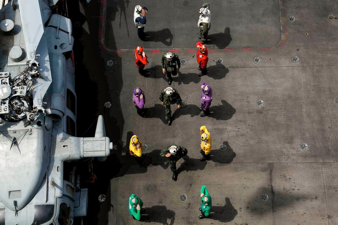 Sailors wearing white, red, purple, yellow and green uniforms, seen from overhead, stand in two lines and salute Thai military officials.