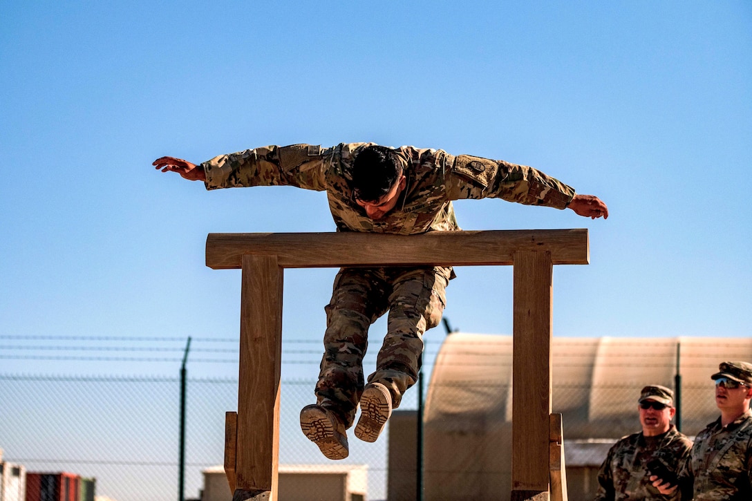 A soldier holds his arms out and leans over a raised horizontal wood plank as two soldiers watch.