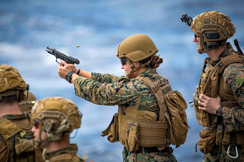 A Marine fires a gun as others watch, framed by a blue ocean.