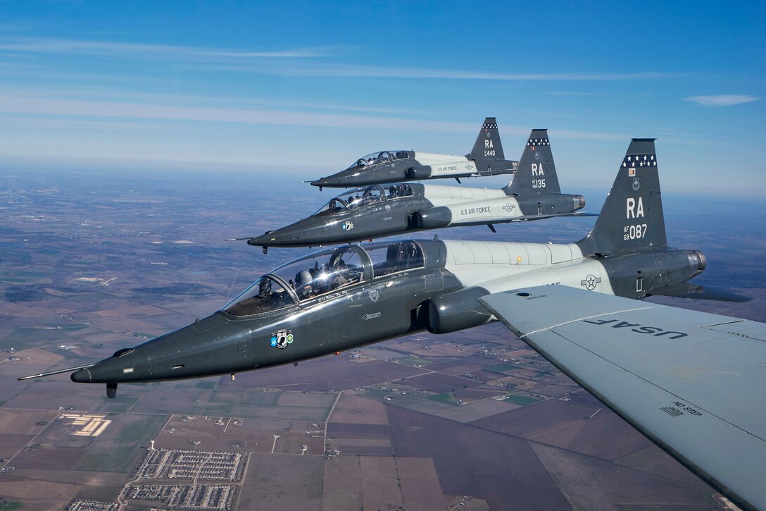 Three aircraft fly in formation in blue sky with a fourth, a wing of which is visible.