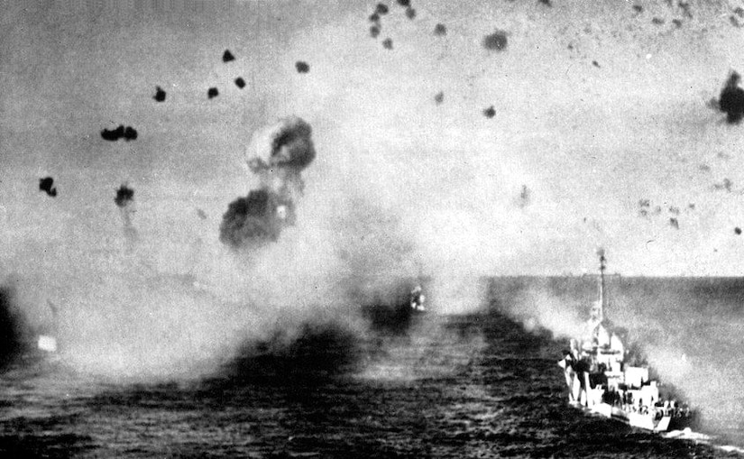 Ships under attack with debris and smoke in the air in a black and white photo.
