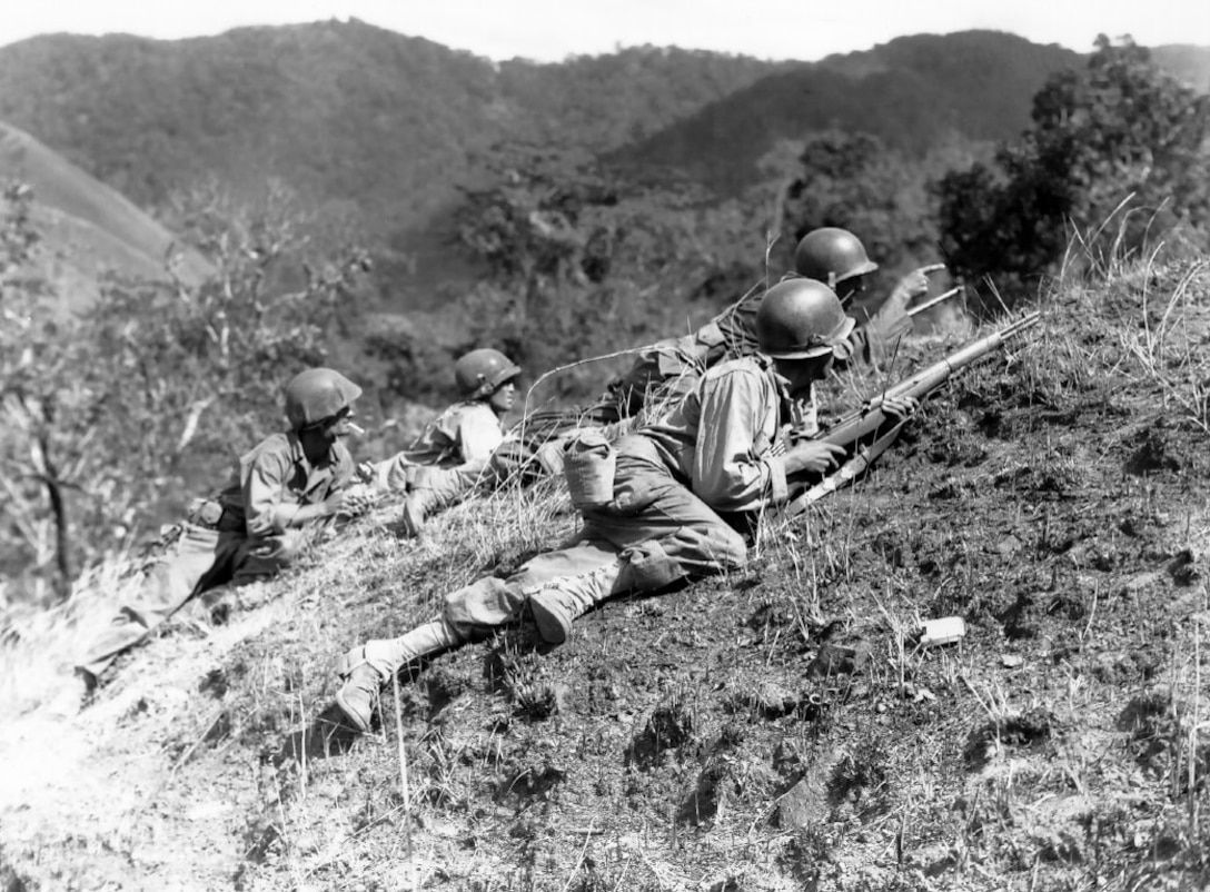 Soldiers crawl up a hill with mountains in the background.