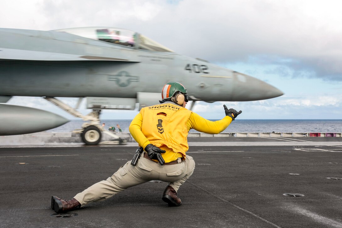 A sailor in a yellow shirt, shown from behind, crouches and gestures to an aircraft in front of him on a ship deck.