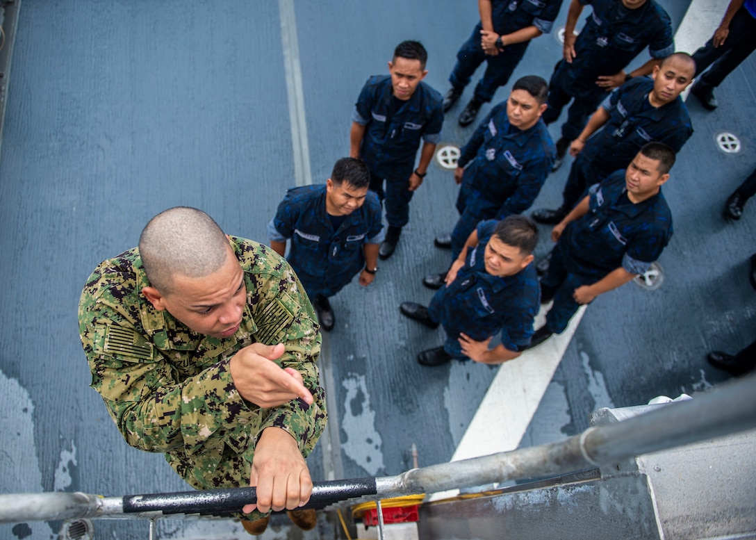 A sailor in camouflage climbs a ladder while sailors in blue uniforms watch from the deck below.
