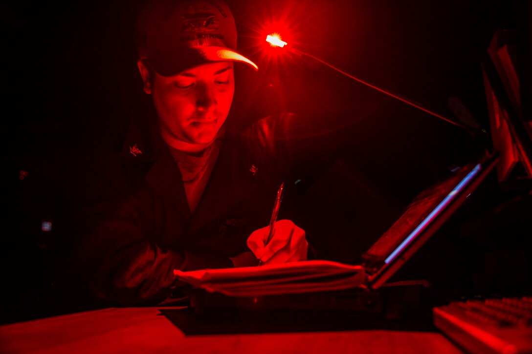 A sailor writes in a book in a dark area illuminated by red light.