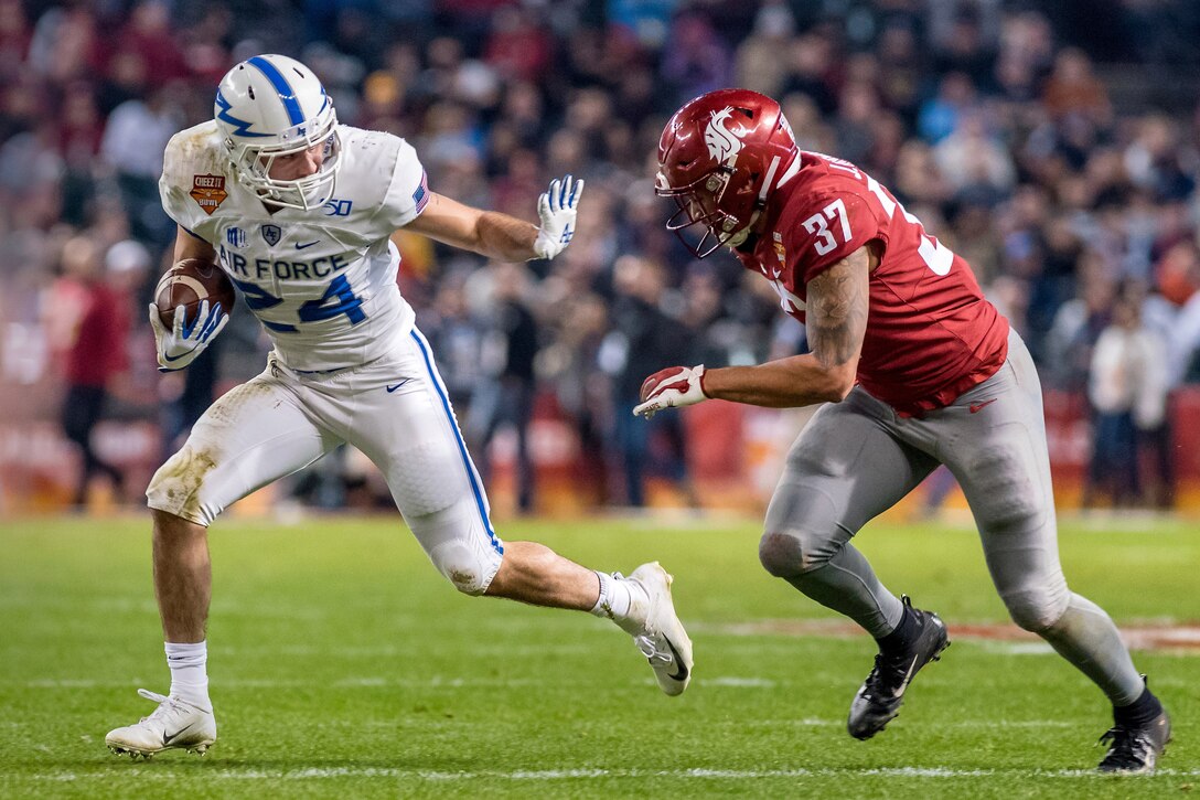 A football player holds his arm out in a blocking gesture to another player running behind him.