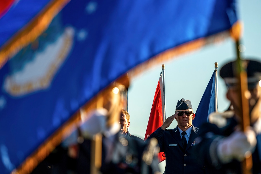 An airman salutes in the background, framed by a blue flag held by an honor guard member in the foreground.
