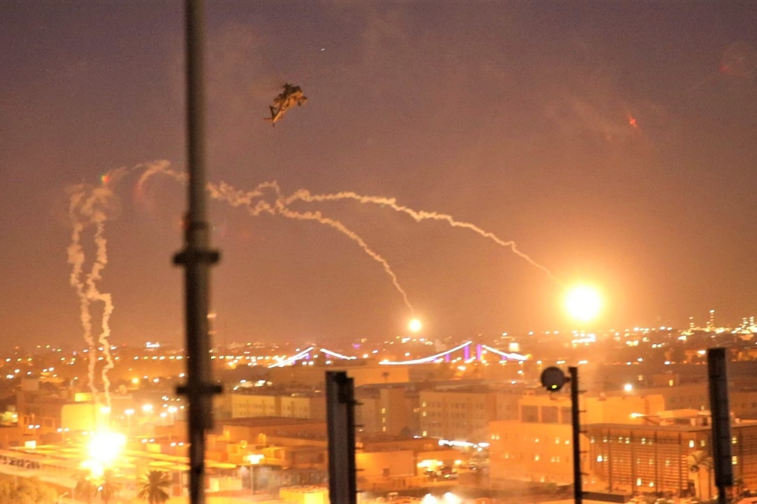 A helicopter flies above an urban area as flares illuminate the sky.
