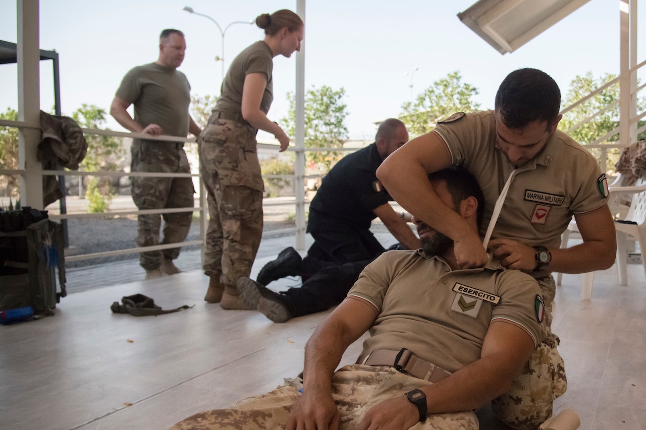 A man leans against another man as gauze is applied to his shoulder.