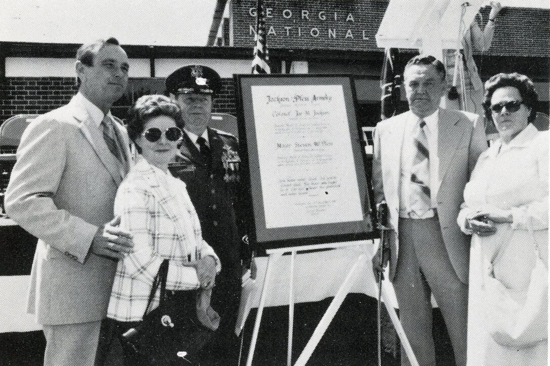 Five people stand in front of a plaque in front of an armory.