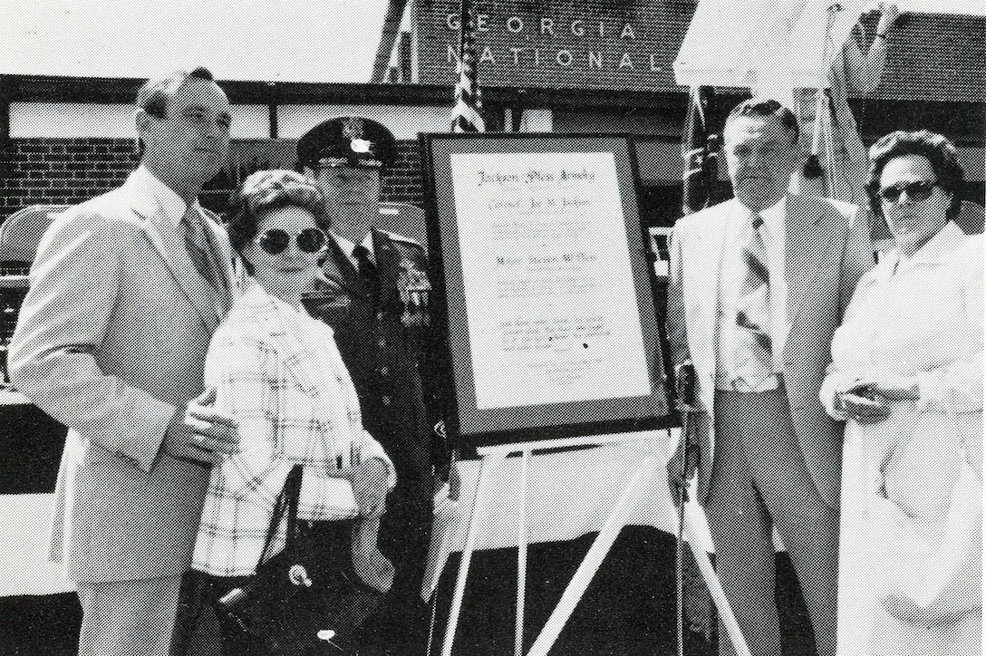 Five people stand in front of a plaque in front of an armory.