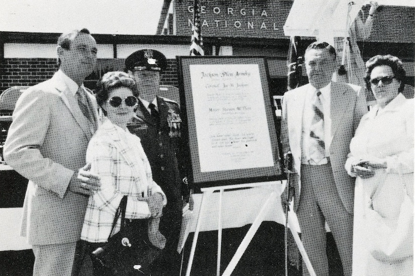 Five people stand in front of a plaque in front of an armory.
