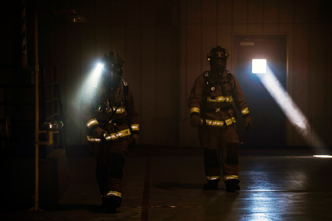 Two firefighters walk through a large smokey room.