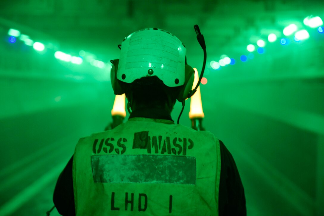 A sailor uses yellow wands to guide a landing craft in the well deck of a military ship.