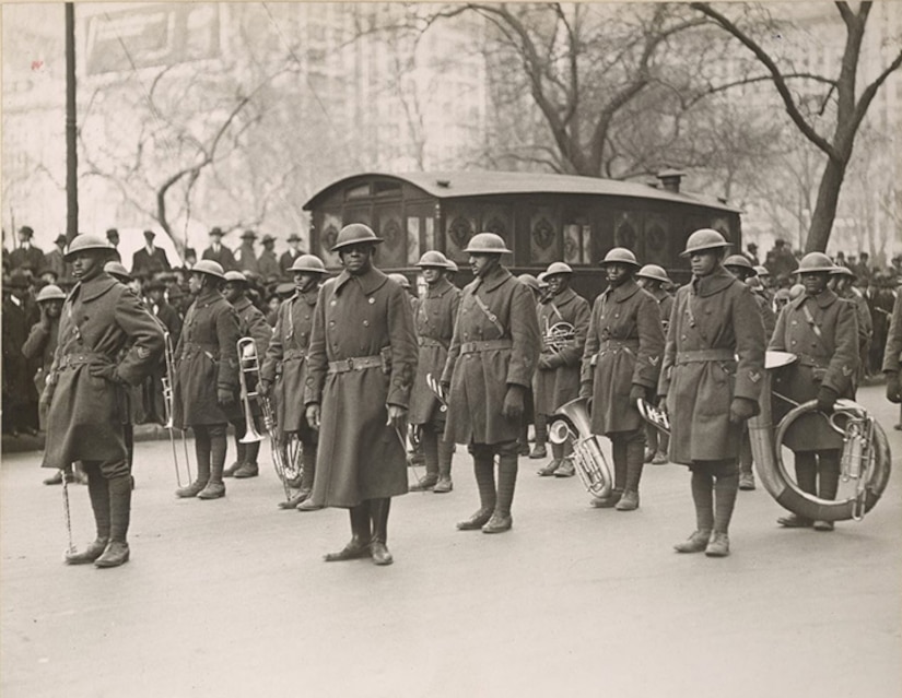 A little over a dozen men in early 20th-century military uniforms are standing in formation outside, holding wind instruments. Some of them are looking toward the camera.