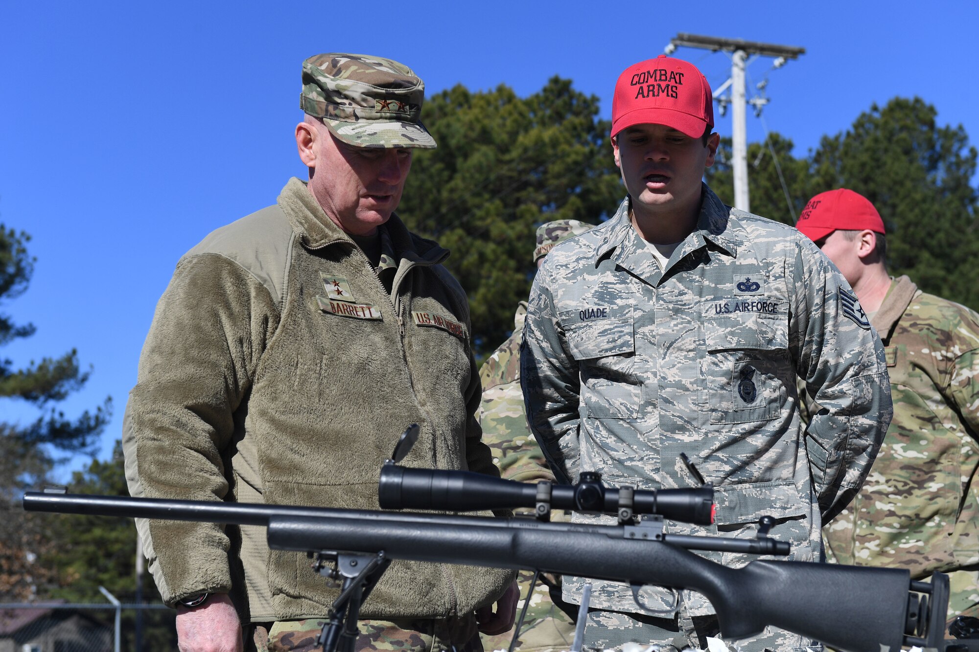 Two men stand in front of a rifle and talk.